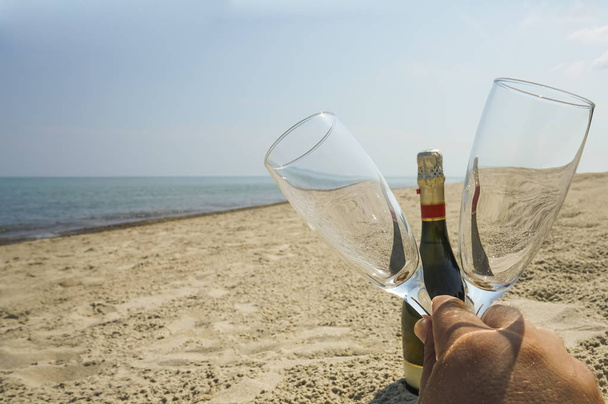 male hand holding two empty clear crystal glasses on one full bottle of champagne. summer sandy beach.  - Photo, image