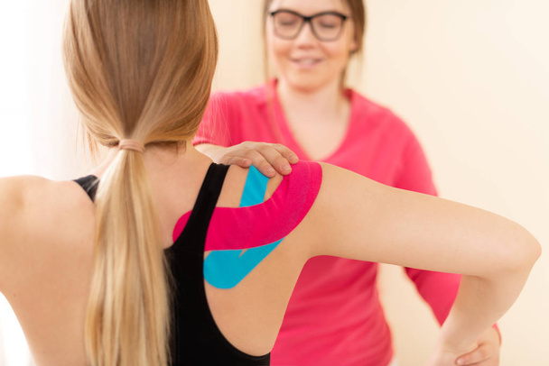 Young female patient wearing kinesio tape on her shoulder exercising with a professional physical therapist. Kinesiology, physical therapy, rehabilitation concept. - Fotó, kép