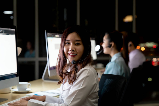 Ázsiai fiatal felnőtt bizalomkezelő nő ügynök headset dolgozik egy call center éjjel Környezet kollégájával csapata, mint az ügyfélszolgálat és a technikai támogatás. Használata késő este Keményen dolgozik 24 Hr. 7 napos koncepció. - Fotó, kép