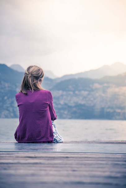 Young girl is sitting on a footbridge and enjoys the view over the lake and the mountains - Φωτογραφία, εικόνα