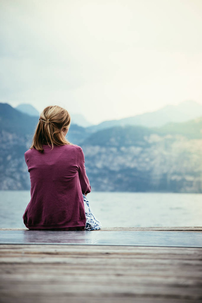 Young girl is sitting on a footbridge and enjoys the view over the lake and the mountains - Photo, Image
