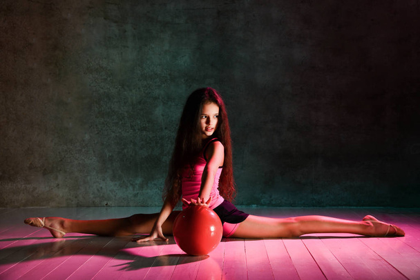 Flexible teen girl with long hair teen doing rhythmic gymnastic exercises with red ball sitting on floor stretching - Foto, Bild