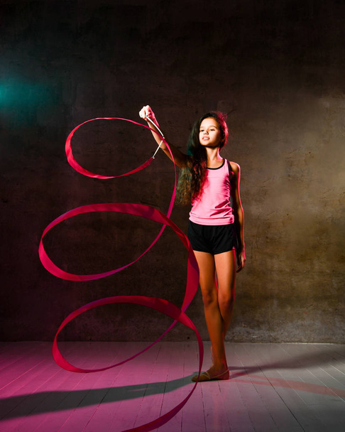 Athletic teen with long curly hair girl in sports uniform is doing rhythmic gymnastic exercises with tape, doing spiral - Valokuva, kuva