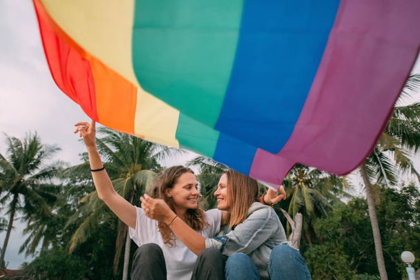 Twee vrouwen met regenboogvlag op het strand op een palmachtergrond - Foto, afbeelding