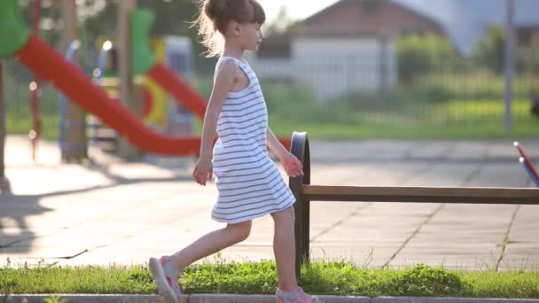 Trauriges Mädchen, das auf einer Bank auf dem Sommerspielplatz im Kindergartenhof auf seine Mutter wartet. - Filmmaterial, Video