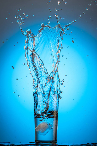 large splash and stream in a glass with clear water on a blue cyan background from an ice cube - Foto, immagini