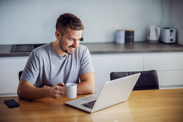 Молодой улыбающийся мужчина одет непринужденно сидя за обеденным столом, держа кружку с утренним кофе и глядя на ноутбук. Он посещает сайты онлайн знакомств
. - Фото, изображение