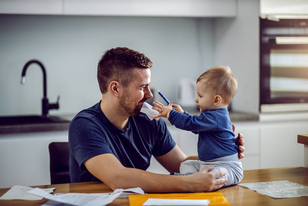 Young proud dad sitting at dining table, having bill in mouth and holding his son. Toddler is sitting on table and helping his dad to fill in bill. - 写真・画像