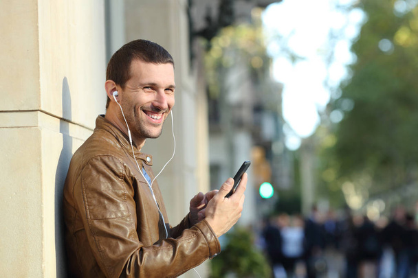 Happy man wearing earphones holding phone looks at you - Foto, afbeelding