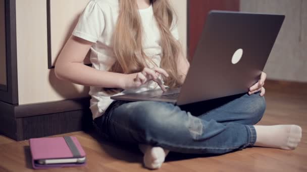 A smart little girl of seven years old in white headphones with a laptop in her hands is pushing on the floor in her room. The young generation on the Internet and IT technology - Felvétel, videó