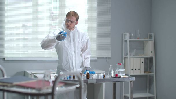 Hombre en ropa de trabajo protectora, gafas y guantes médicos revisa tubos en el laboratorio
 - Foto, Imagen