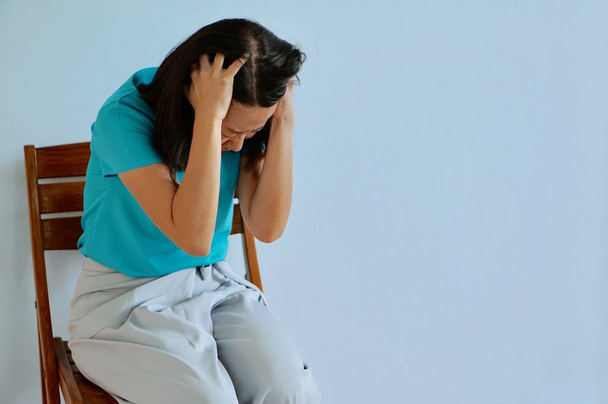 A young asian woman in turquoise color sitting on a chair getting headache, blue background - Photo, Image