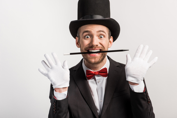 excited magician in suit and hat holding wand in teeth, isolated on grey - Fotoğraf, Görsel