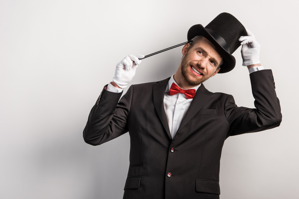 задумчивый улыбающийся фокусник в костюме и шляпе с палочкой, изолированный на сером
 - Фото, изображение