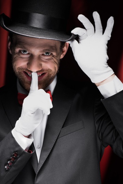 scary smiling magician winking and showing silence symbol in circus with red curtains - Фото, изображение