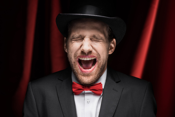 ijesztő érzelmi bűvész csukott szemmel és nyitott szájjal cirkuszban vörös függönyökkel - Fotó, kép