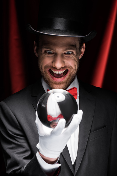 vzrušený děsivý kouzelník při pohledu na kouzelný míč v cirkusu s červenými závěsy - Fotografie, Obrázek