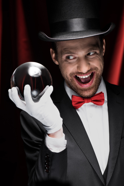 vzrušený děsivý kouzelník drží magický míč v cirkusu s červenými závěsy - Fotografie, Obrázek