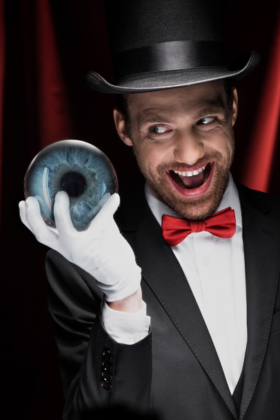 vzrušený děsivý kouzelník drží modrý magický míč v cirkusu s červenými závěsy - Fotografie, Obrázek