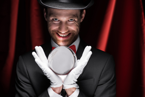 przerażający uśmiechnięty magik trzymając magiczną piłkę w cyrku z czerwonymi zasłonami - Zdjęcie, obraz