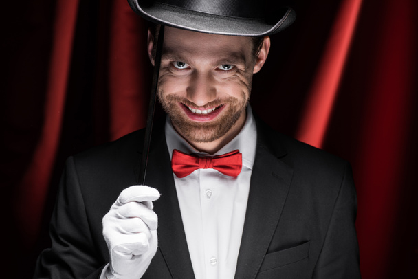 glimlachende enge goochelaar in pak en hoed stok in circus met rode gordijnen - Foto, afbeelding