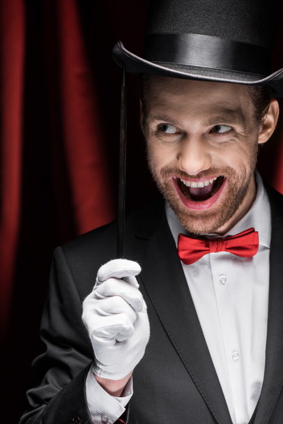 mágico animado com boca aberta segurando varinha no circo com cortinas vermelhas
 - Foto, Imagem