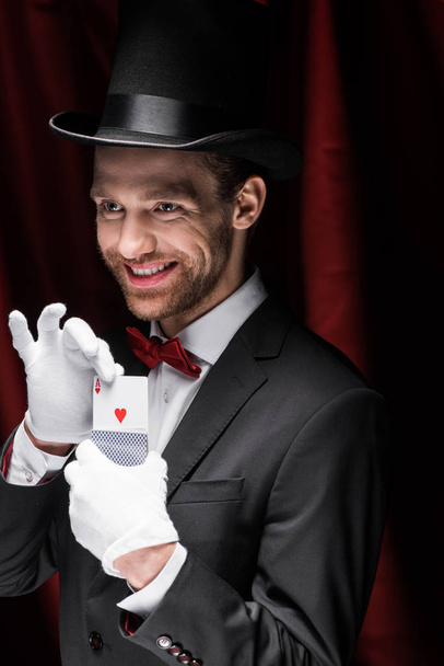 smiling magician holding playing cards in circus with red curtains - Фото, зображення