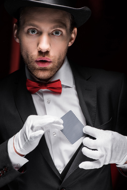 surprised magician holding playing card in circus with red curtains - Fotoğraf, Görsel