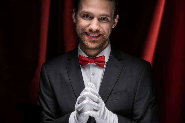 uśmiechnięty dżentelmen w garniturze, muszce i rękawiczki w cyrku z czerwonymi zasłonami - Zdjęcie, obraz