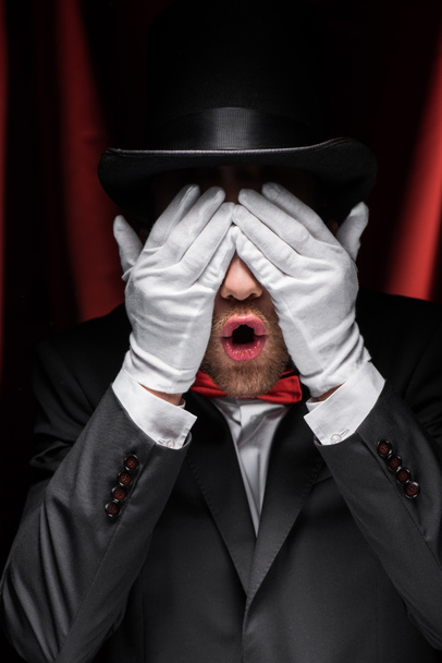 rémült bűvész becsukja a szemét kesztyűvel a cirkuszban piros függönyökkel - Fotó, kép