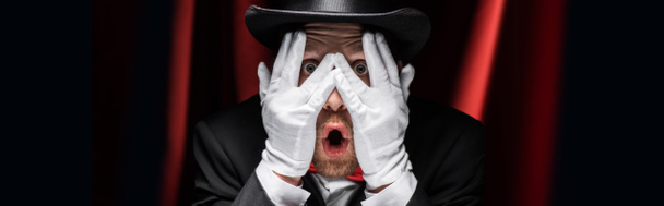 panoramic shot of frightened magician closing eyes with hands in gloves in circus with red curtains - Fotoğraf, Görsel
