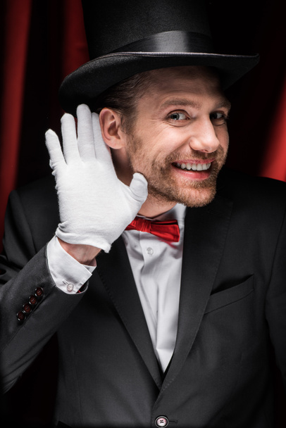 smiling magician gesturing and listening in circus with red curtains - Фото, изображение