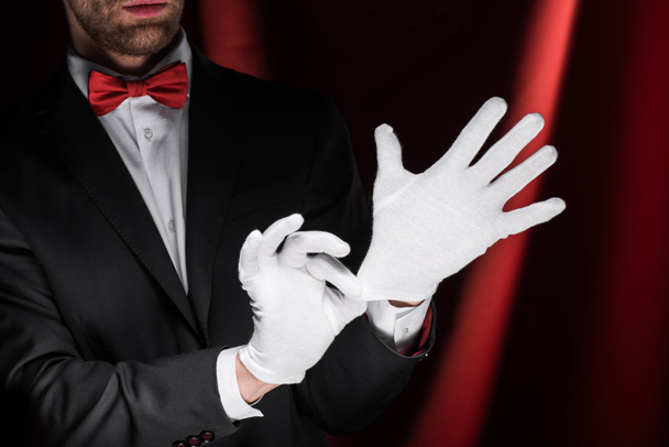 bijgesneden zicht van goochelaar dragen handschoenen in circus met rode gordijnen - Foto, afbeelding