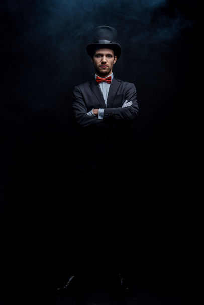 magician in suit and hat standing with crossed arms in dark smoky room - Photo, Image