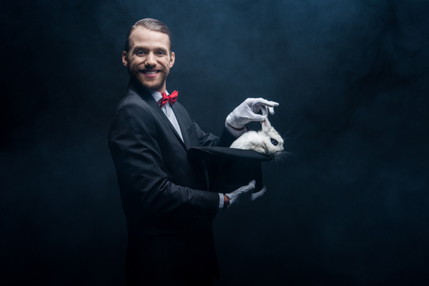 fröhlicher Zauberer im Anzug zeigt Trick mit weißem Kaninchen im Hut, dunkler Raum mit Rauch - Foto, Bild