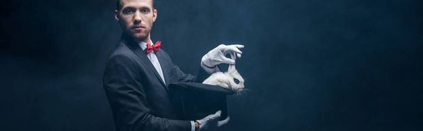 Panoramaaufnahme eines jungen Zauberers im Anzug, der Trick mit weißem Kaninchen im Hut zeigt, dunkler Raum mit Rauch - Foto, Bild