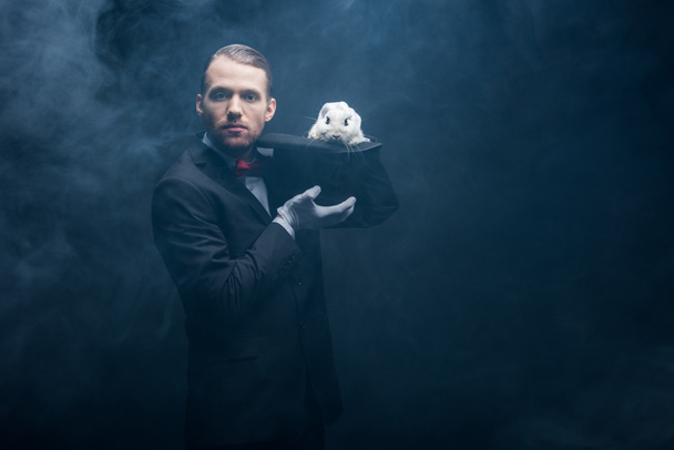 professional magician in suit showing trick with white rabbit in hat, dark room with smoke - Photo, Image