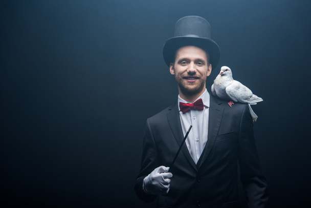 Taube sitzt auf der Schulter eines fröhlichen Zauberers mit Hut und Zauberstab im dunklen Raum mit Rauch  - Foto, Bild