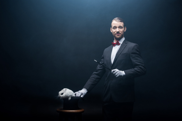 happy magician in suit showing trick with wand and white rabbit in hat, dark room with smoke - Photo, image