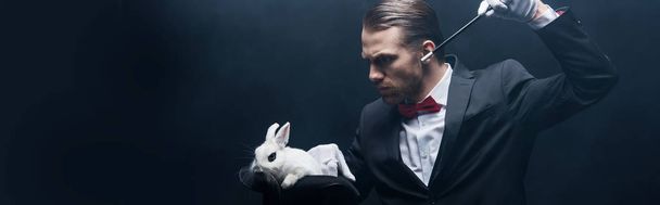 panoramic shot of concentrated young magician in suit showing trick with wand and white rabbit in hat, dark room with smoke - Fotoğraf, Görsel