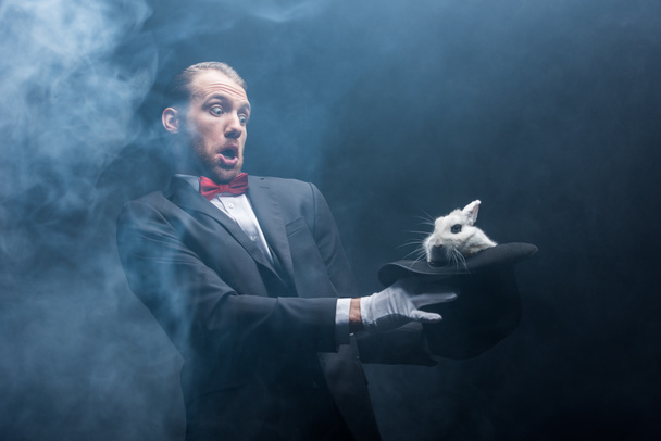 frightened magician holding white rabbit in hat, dark room with smoke - Photo, Image