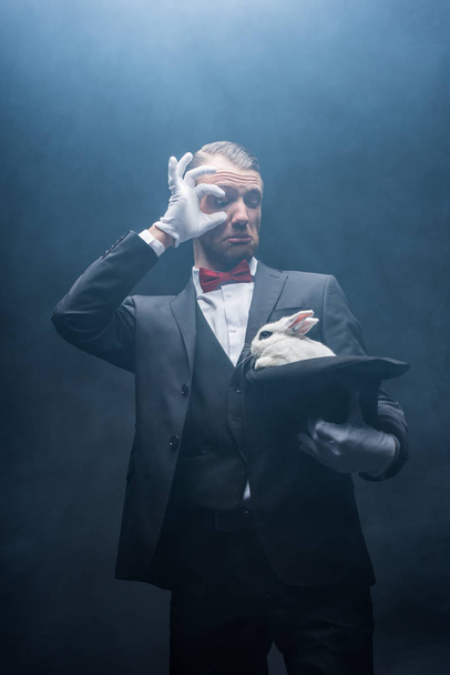 surprised magician looking at white rabbit in hat, dark room with smoke - Valokuva, kuva