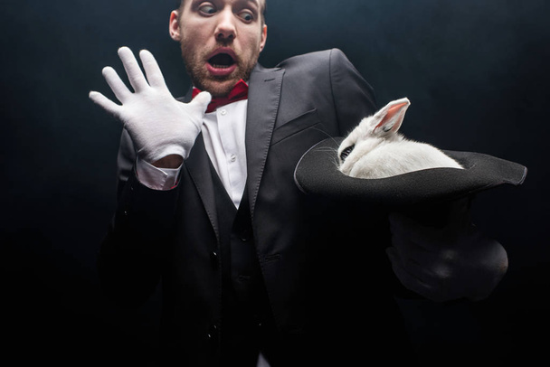 scared magician holding white rabbit in hat, isolated on black - Photo, Image