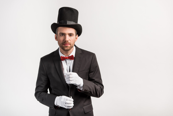 handsome magician in suit and hat holding wand, isolated on grey - Fotó, kép