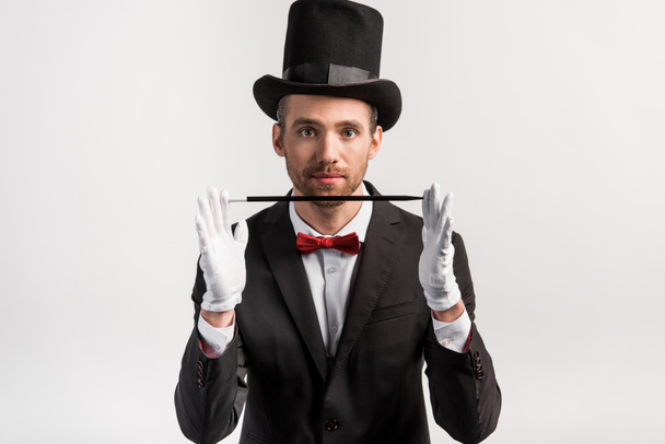 mladý kouzelník v obleku a klobouku držící hůlku, izolovaný na šedi - Fotografie, Obrázek