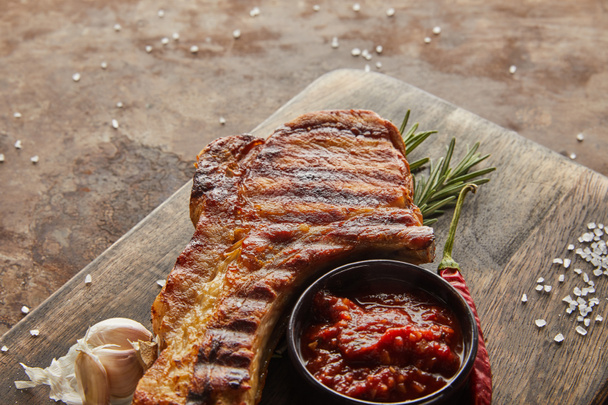 Close up view of tasty steak with rosemary, garlic and tomato sauce on chopping board on stone surface - Fotografie, Obrázek
