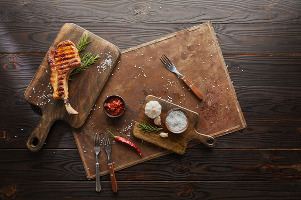 Draufsicht auf leckeres Steak auf Schneidebrett mit Chilisoße, Rosmarin und Salz auf Steinplatte auf Holzgrund - Foto, Bild