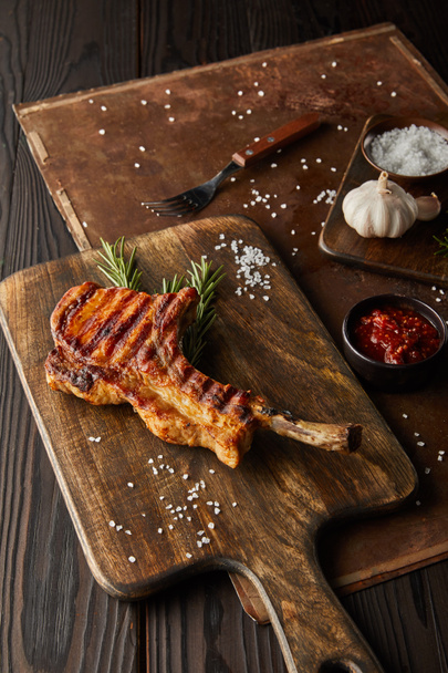 Selective focus of gourmet ribeye steak on cutting board with salt, tomato sauce and garlic on stone board on wooden background - Photo, Image