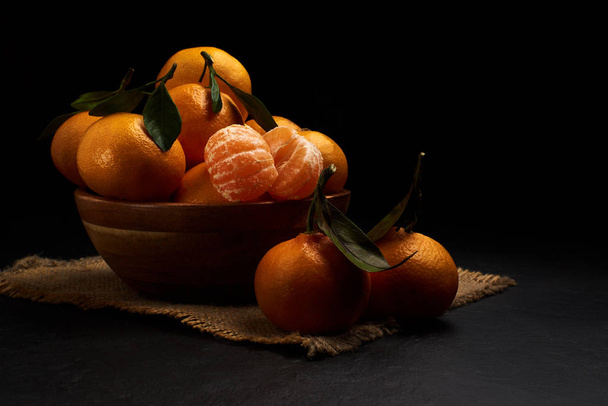 Ripe mandarins in wooden bowl on table with sackcloth, black background. - Photo, image