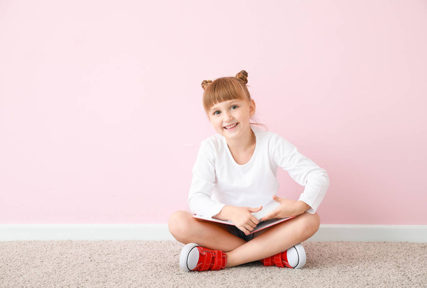 Χαριτωμένο κοριτσάκι με το βιβλίο κάθεται κοντά στον τοίχο χρώμα - Φωτογραφία, εικόνα